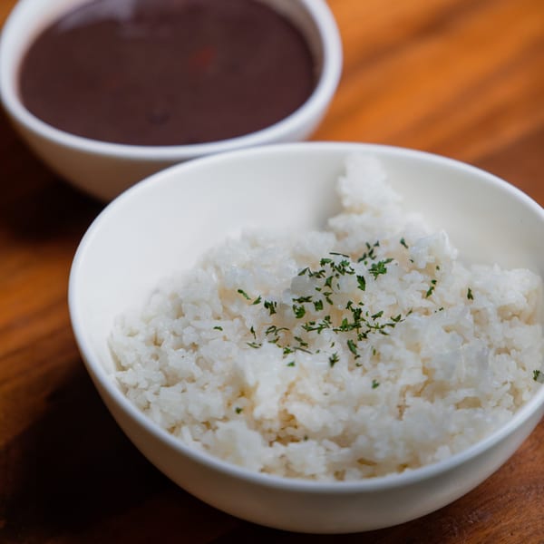 A white bowl of fluffy white rice topped with parsley, with a bowl of dark beans behind it.