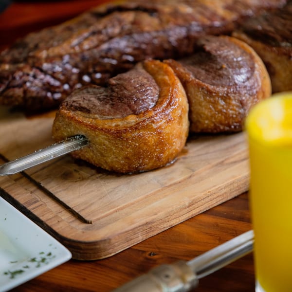 Brazilian picanha steak on a skewer, resting on a wooden board with a drink beside it.